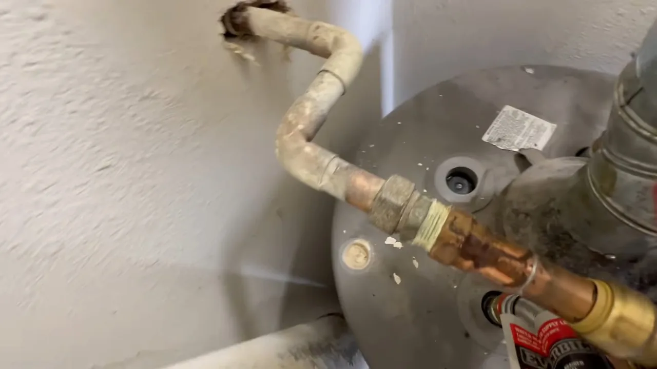 Aged supply pipe and a brass union connected to the top of a grey water heater against a textured wall.