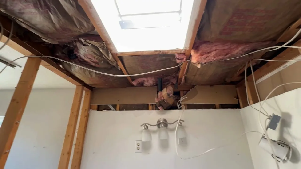 Bathroom ceiling cut open to bare joists and pink insulation above a vanity light, with a skylight opening.