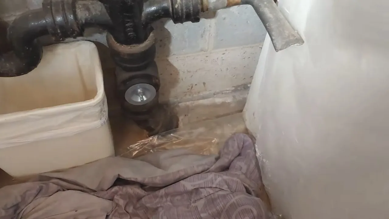 Black cast iron drain trap and pipe at a bathroom floor, with a plastic bucket and a cloth beside it.