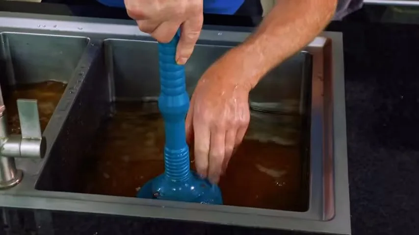 Hands working a blue plunger in a double kitchen sink full of brown water.