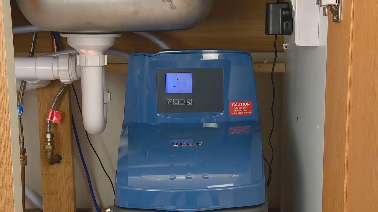 Blue water softener with a digital display standing under a kitchen sink, with the drain trap above it.