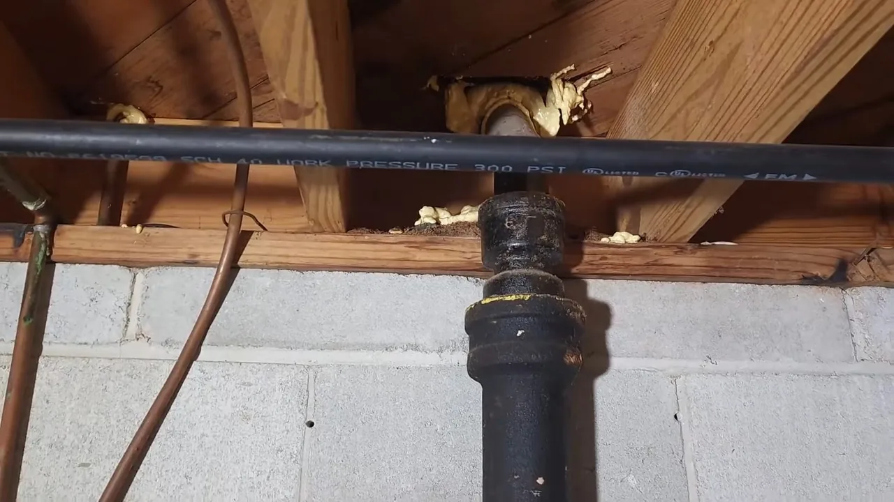 Black cast iron stack with a black plastic line and a copper pipe running past it beneath floor joists, above a concrete block wall.