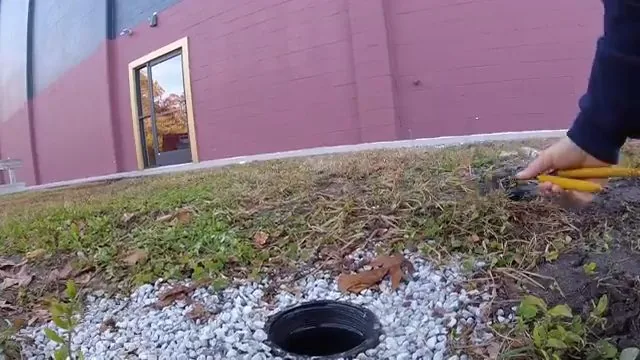 Round cleanout set in gravel beside a lawn, with a hand holding a yellow tool at its edge.