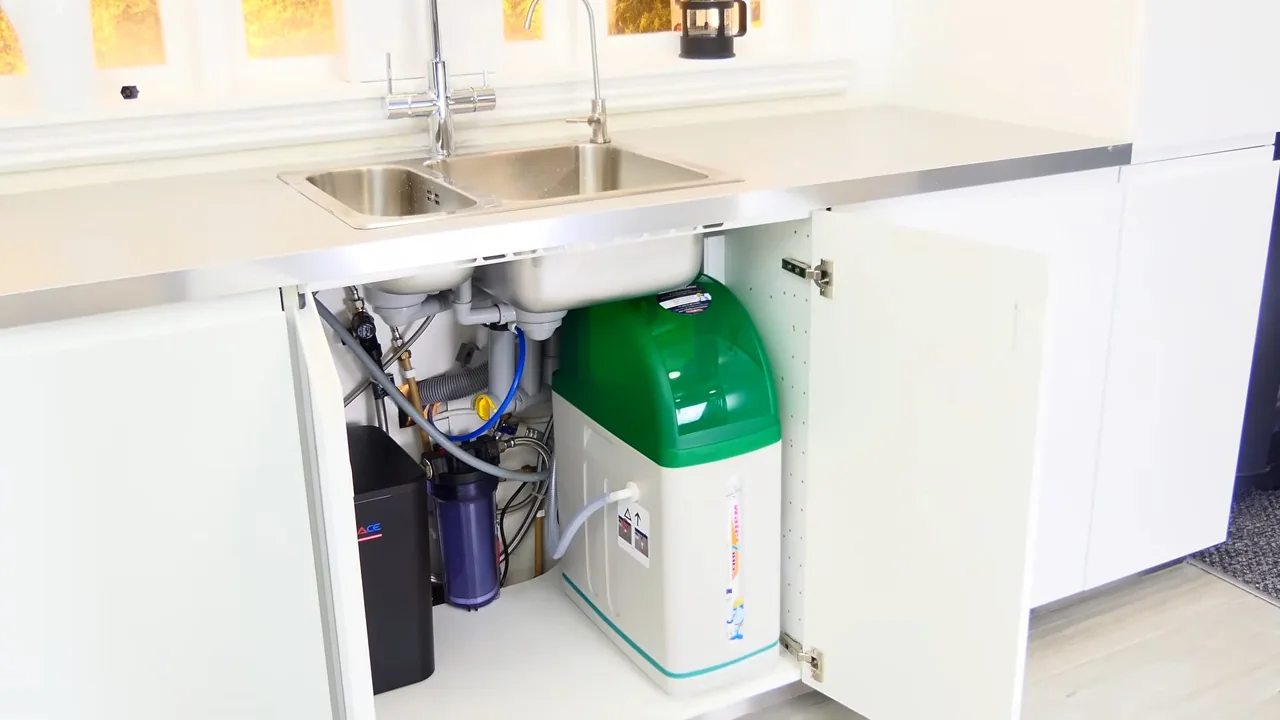 Compact green and white water softener standing in an open cabinet beneath a double kitchen sink.