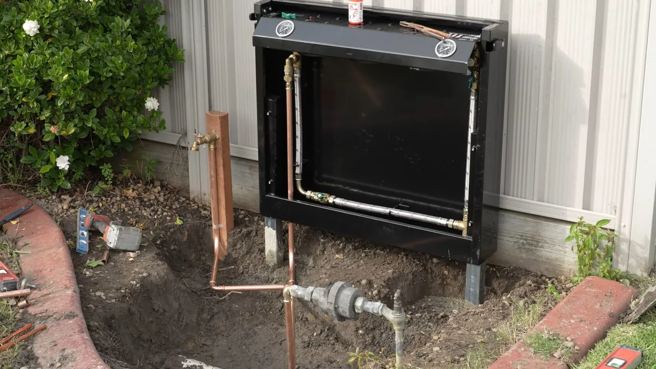 Copper supply line with shutoff valves and a water meter in a dug-out bed beside an exterior wall, under a black frame.