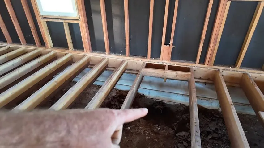Hand pointing toward bare soil visible between exposed floor joists, with open wall framing above.