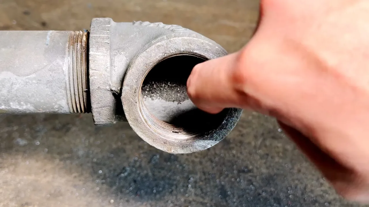 Finger inside the threaded open end of a galvanized pipe elbow lying on a rough surface.