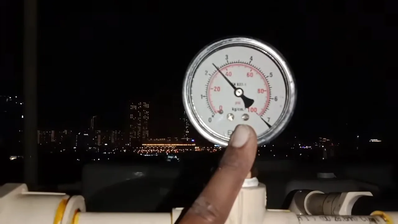 Finger touching a round pressure gauge mounted above white pipe against a dark night sky.