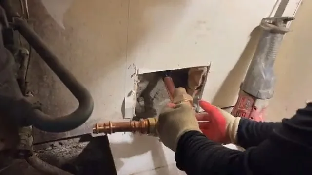 Gloved hands fitting a copper stub-out inside a rectangular opening cut in a wall, with a pipe wrench nearby.
