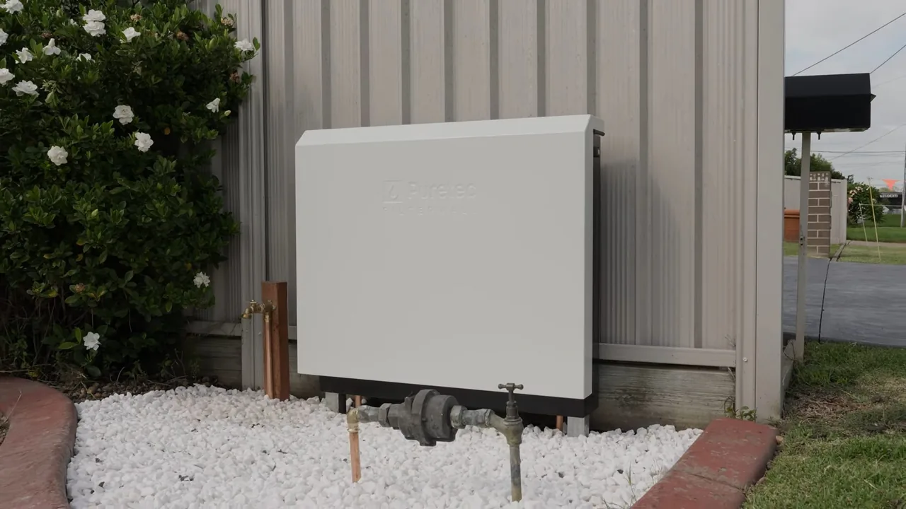 Gray equipment cabinet on white gravel against a house wall, with copper pipe and two shutoff valves at its base.
