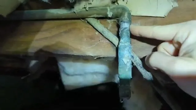 Hand pointing to a green, corroded copper elbow beneath floor framing.