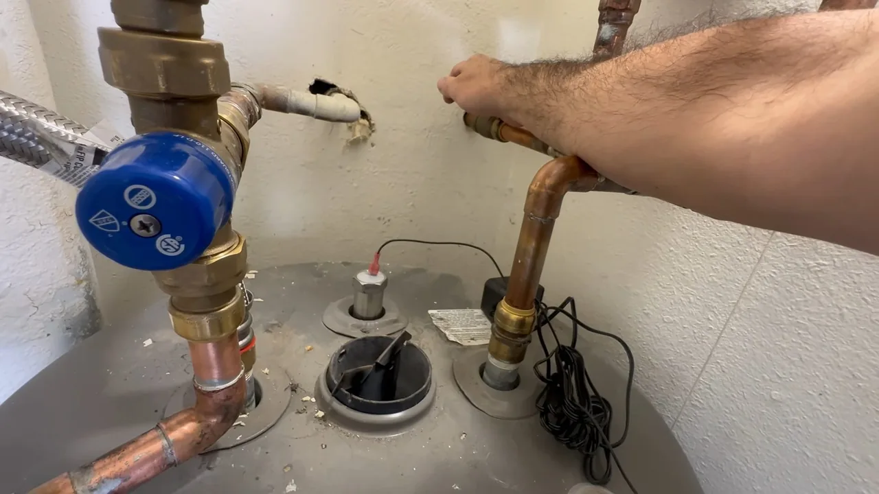 Hand gripping a copper pipe above a grey water heater tank, with a blue valve and brass fittings on the supply lines.