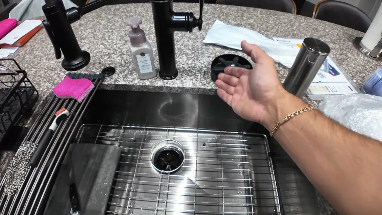 Hand held out over a stainless steel kitchen sink with a drain basket and faucets.