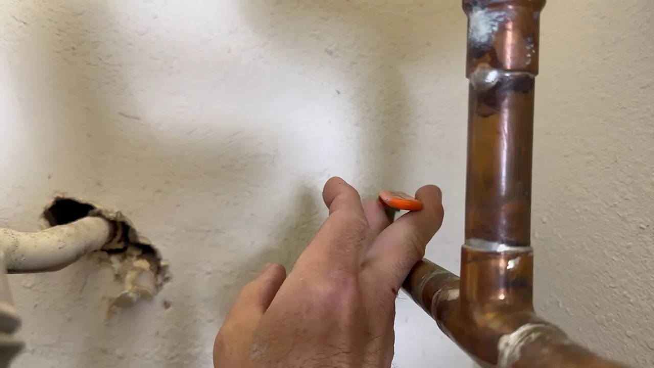 Hand holding a small orange tool against a copper pipe next to a hole cut in a wall.
