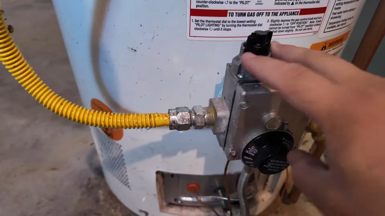 Hand turning the gas control knob on a water heater, with a yellow flexible gas line connected beside it.