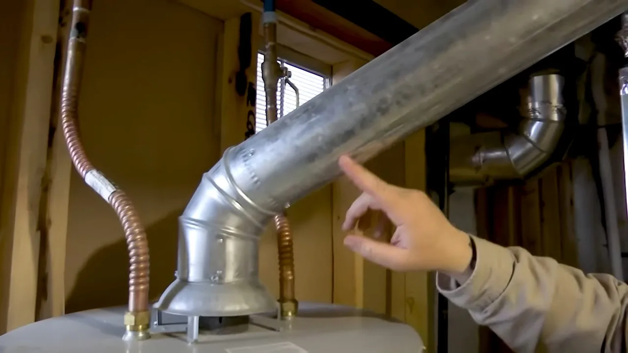 Hand pointing at a galvanised flue elbow rising from a water heater, with flexible connector lines on either side.