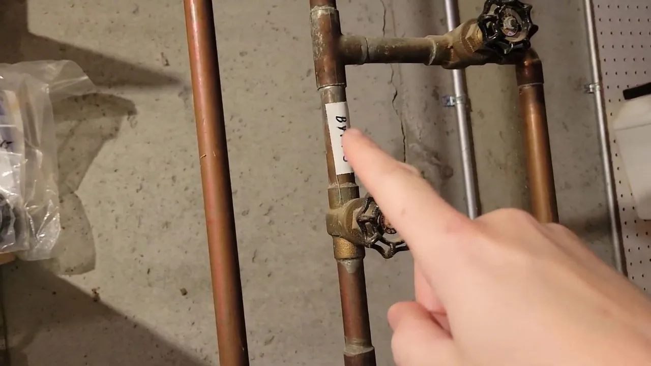 Hand pointing at a brass gate valve on a vertical copper supply line against a concrete wall.