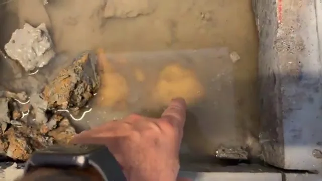 Hand pointing at a wet, discoloured patch on a concrete slab beside broken rubble.