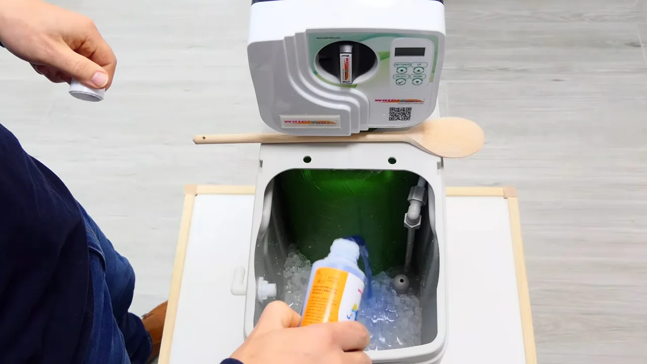 Hand pouring cleaner from an orange bottle into the open brine tank of a white water softener, a wooden spoon resting on the lid.