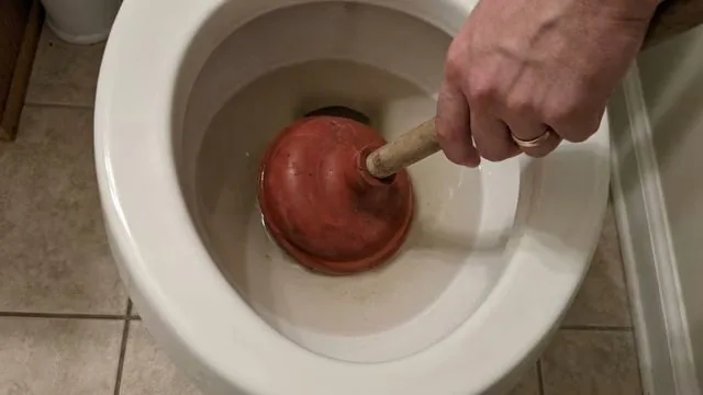 Hand pushing a red plunger into a toilet bowl on a tiled floor.