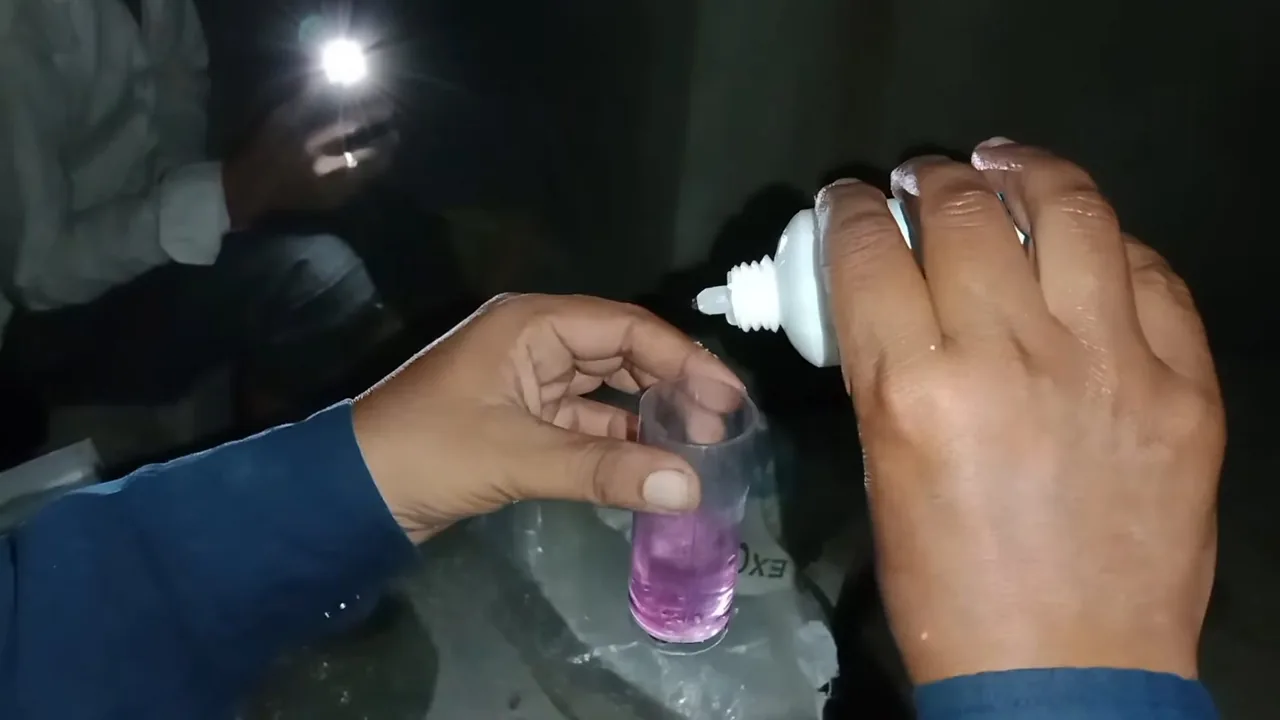 Hand squeezing a dropper bottle over a small glass holding a pink water sample.