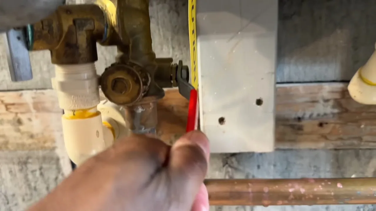 Hand turning the red handle of a brass valve above white CPVC and copper piping.