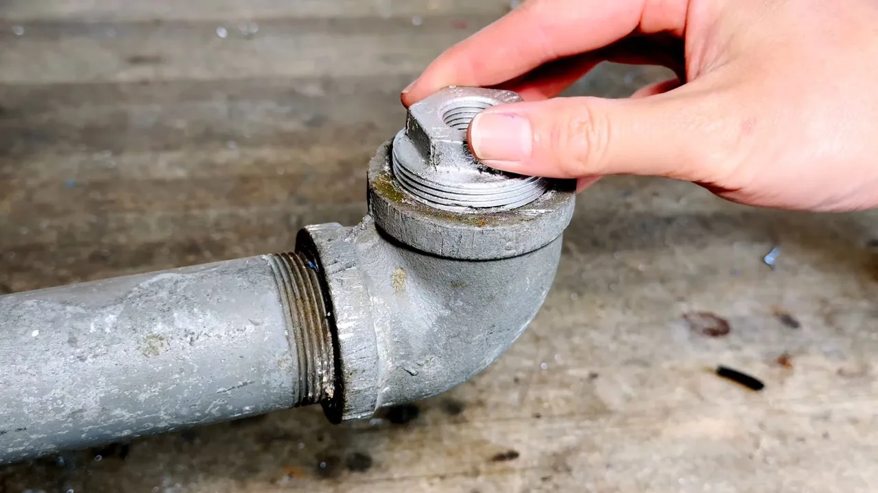 Hand unscrewing the threaded cap on a galvanized steel pipe elbow resting on concrete.