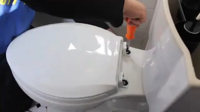Hand using an orange screwdriver at the hinge of a white toilet seat.