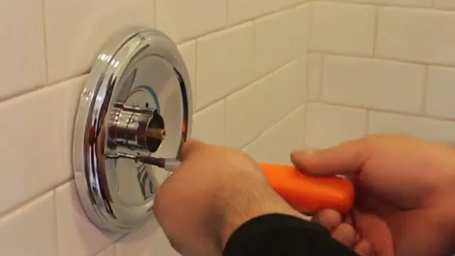 Hand using an orange-handled screwdriver at a chrome shower valve trim plate on a tiled wall.