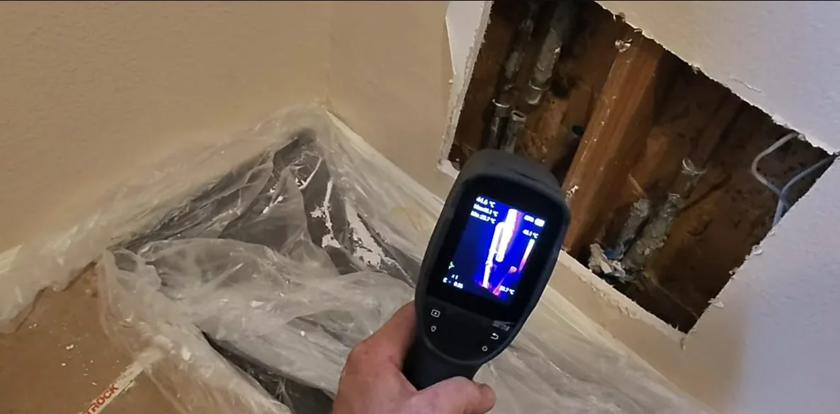 Hand holding a thermal camera toward an opened wall cavity, with a heat pattern on its display.