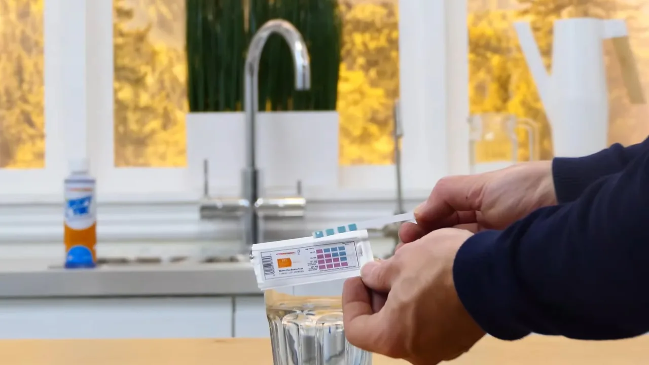 Hands holding a colour test strip over a glass of water beside a kitchen faucet.