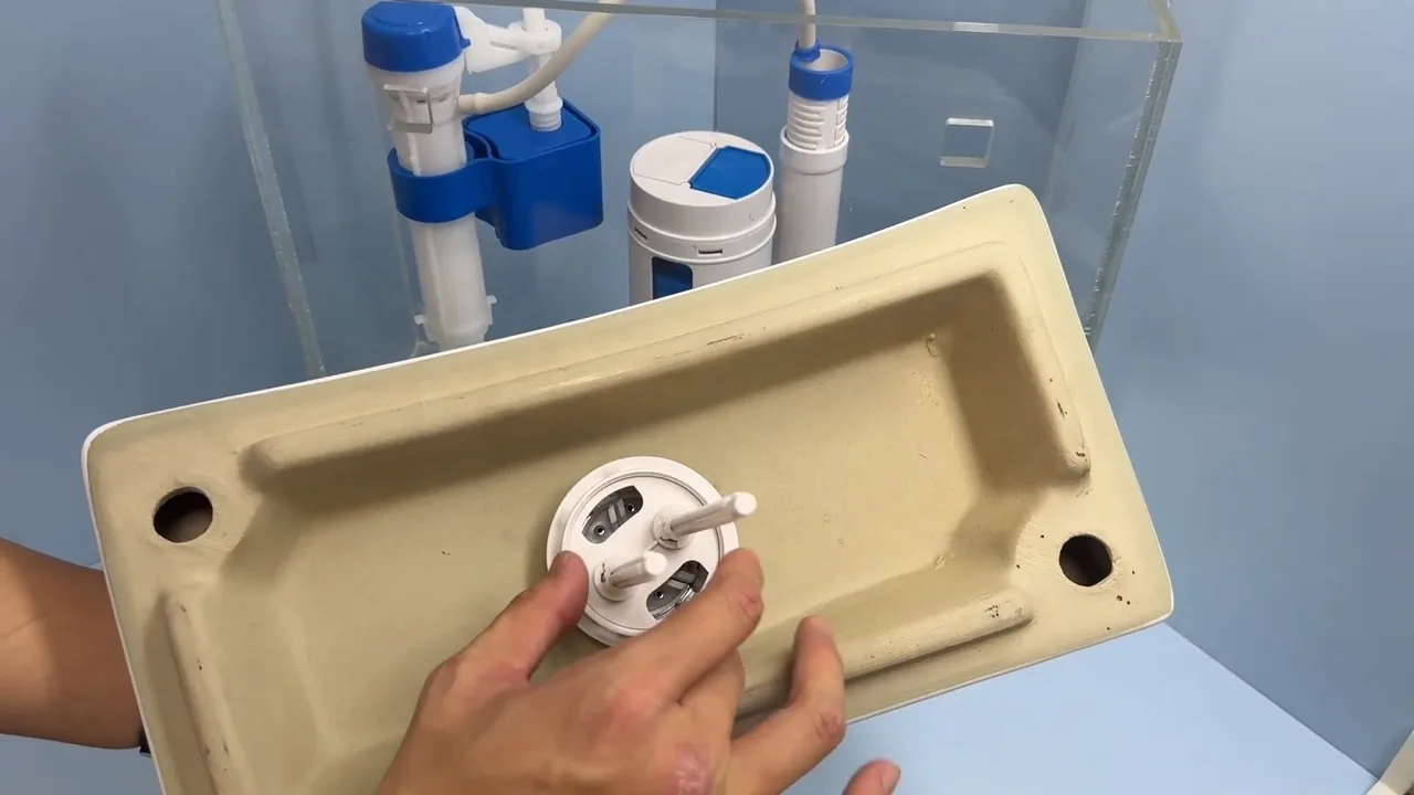Hands holding a toilet tank lid upside down, with a fill valve and other parts in a clear tank behind it.