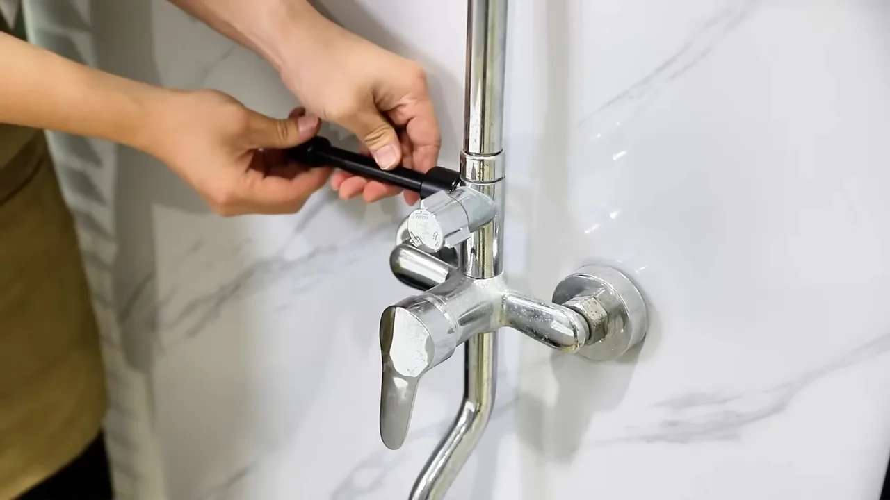 Hands using a small black tool on a chrome wall-mounted shower mixer set against marble-effect tile.
