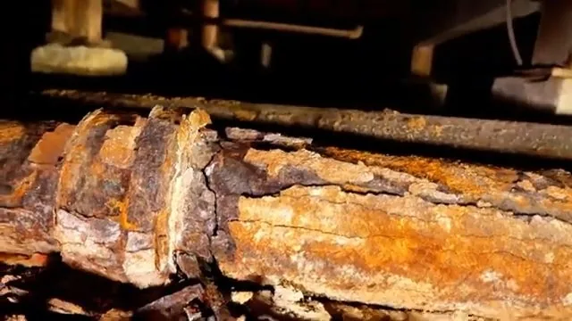 Close view of a heavily rusted cast iron pipe running beneath floor joists, its scale flaking away.