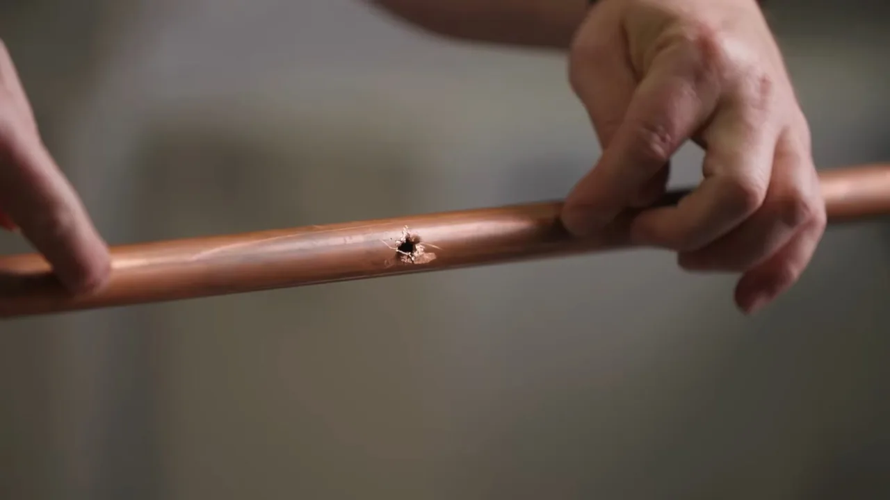 Hands holding a length of copper pipe with a small pinhole in its wall.