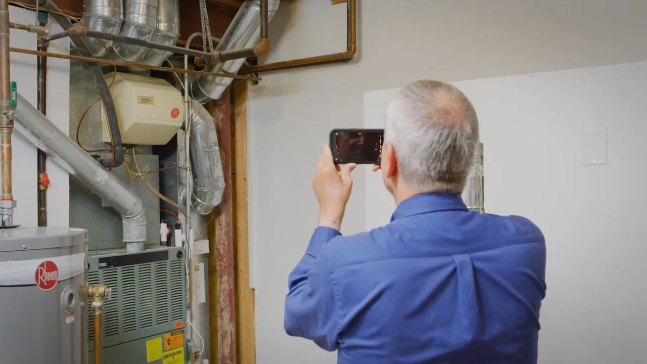 Man in a blue shirt photographing a water heater and the ductwork around it in a utility room.