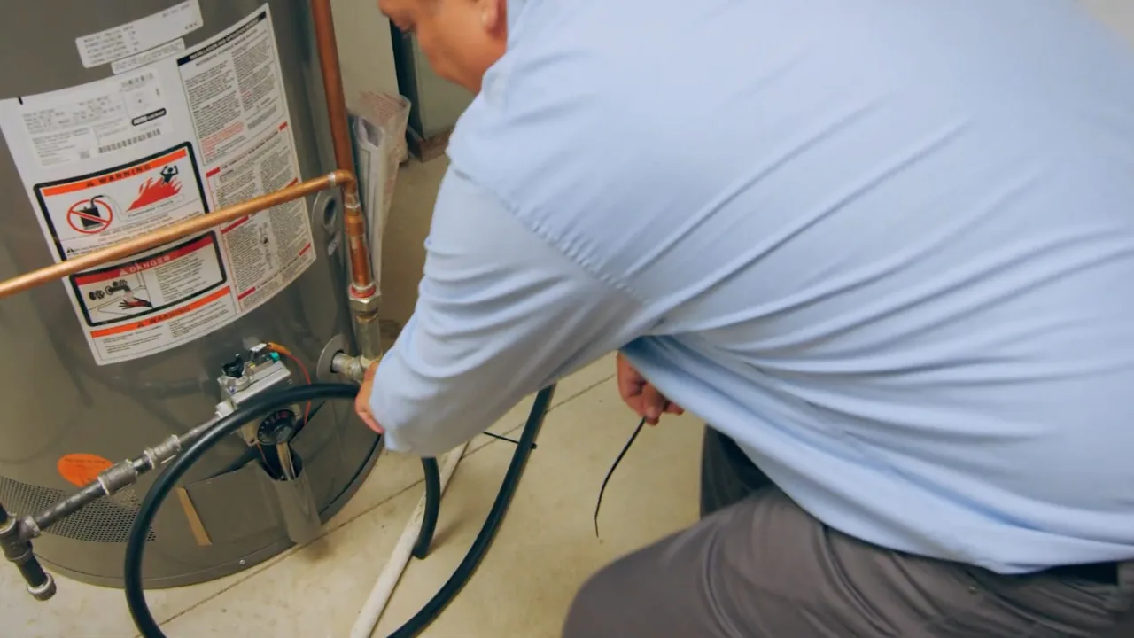 Man in a light blue shirt crouched at a water heater, connecting a dark hose to the lower fitting.