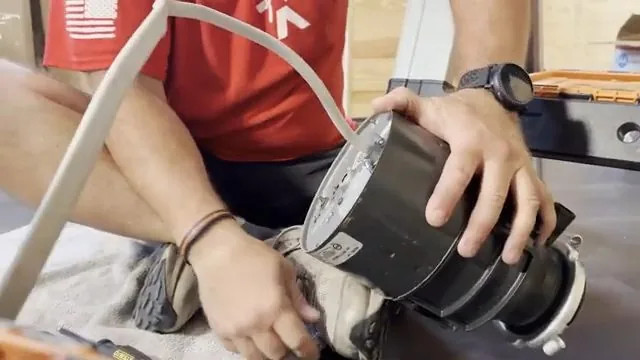 Man in a red shirt holding a garbage disposal with a white cord trailing from its base.
