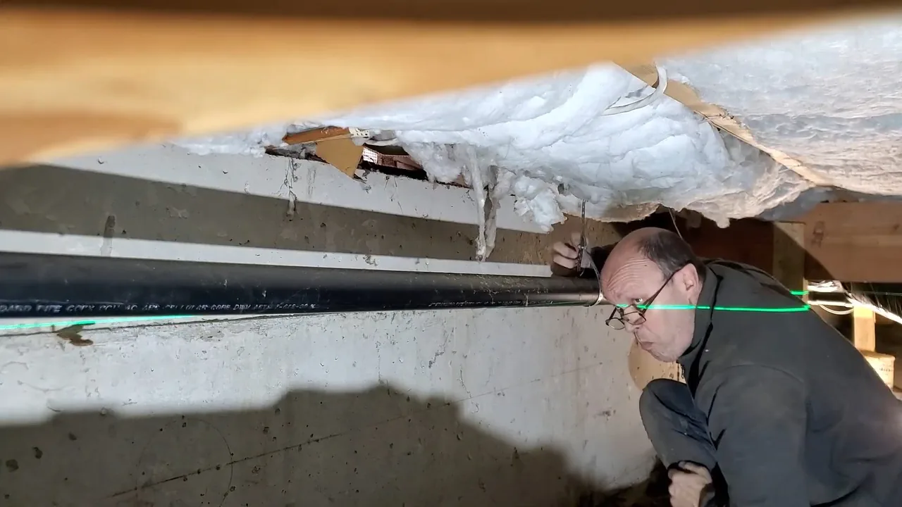 Man crouched under a house floor inspecting a long black pipe run, with a green laser line across the frame.