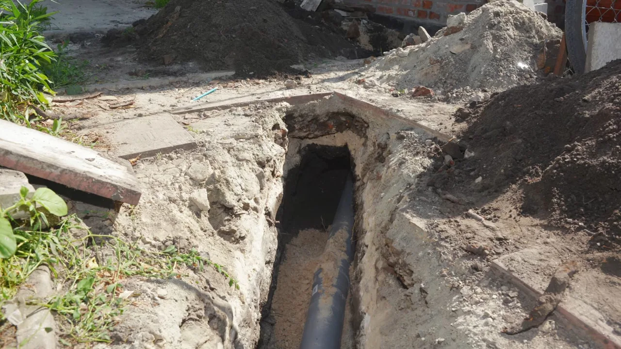 Narrow trench dug beside a patch of lawn with a dark sewer pipe in the bottom and soil piled on the right.