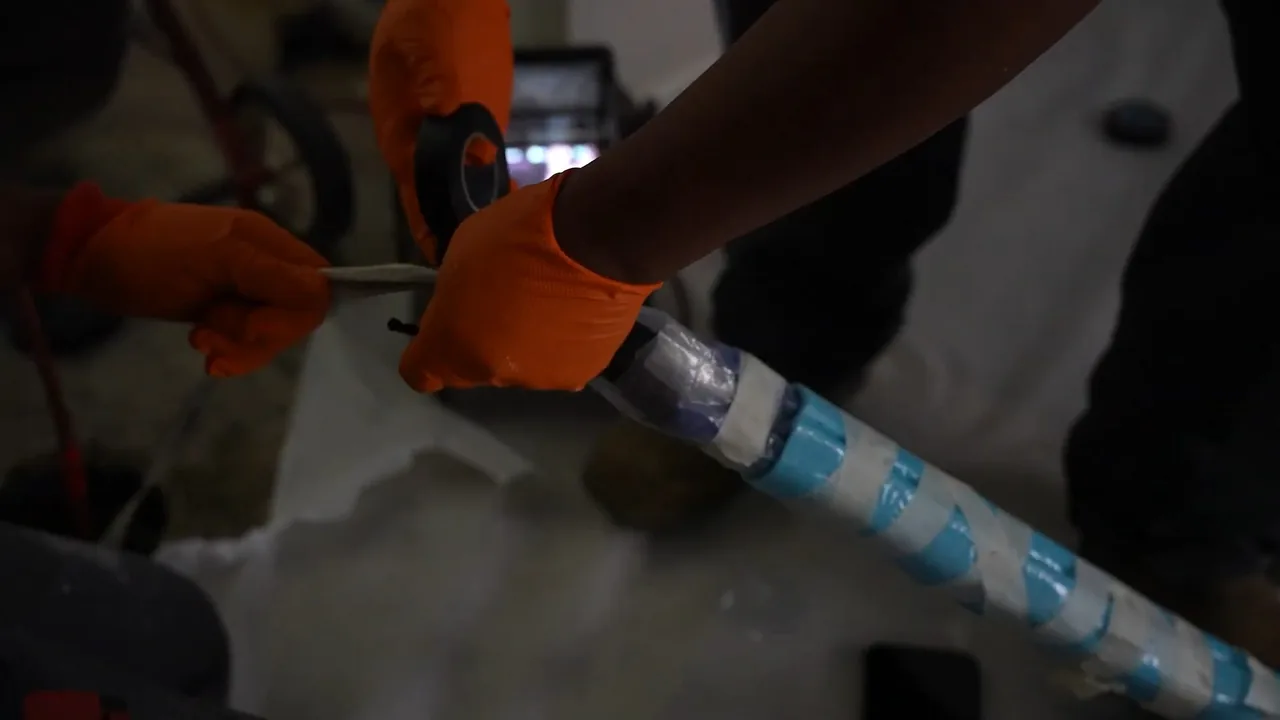 Orange-gloved hands drawing a blue-striped liner tube across a white sheet on the floor.