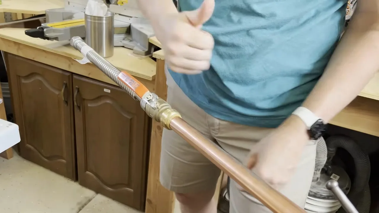 Plumber in a teal shirt giving a thumbs-up while holding a copper supply line beside wooden kitchen cabinets.