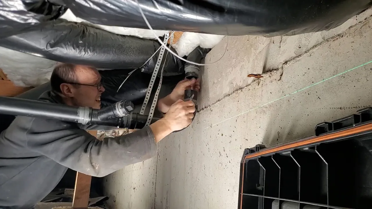Plumber reaching up to a pipe beneath floor joists, with a green laser line along the wall.