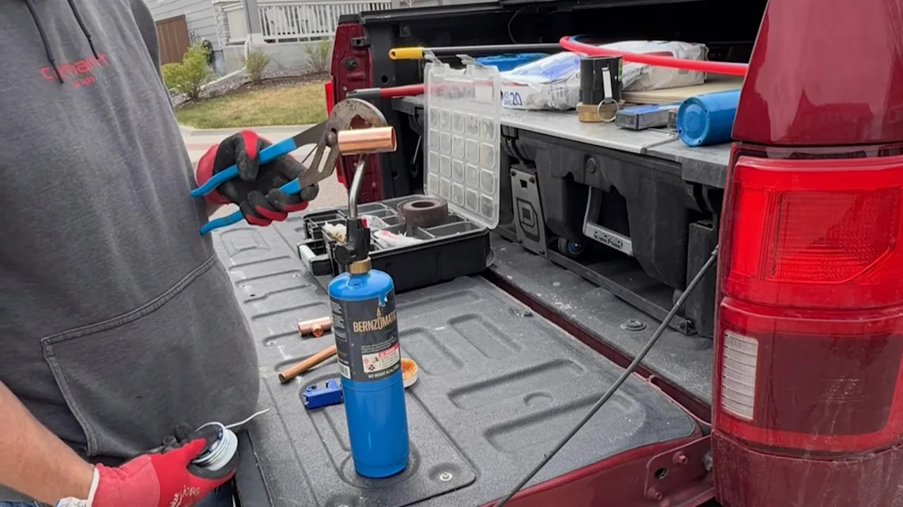 Open truck bed holding a blue propane torch, a tool organizer and pipe-cutting tools, with a worker in a gray shirt holding pliers.