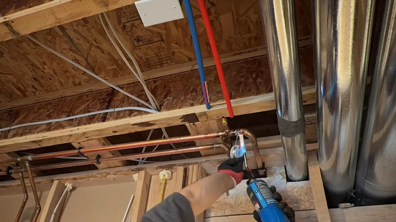 Open ceiling framing with red and blue PEX and copper pipe running between joists, and a hand holding a torch at a joint.