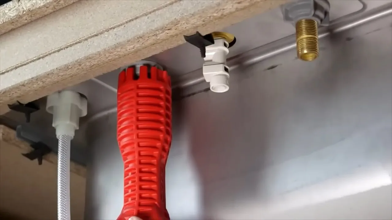 Red basin wrench held under a sink next to a braided supply line and faucet shank fittings.