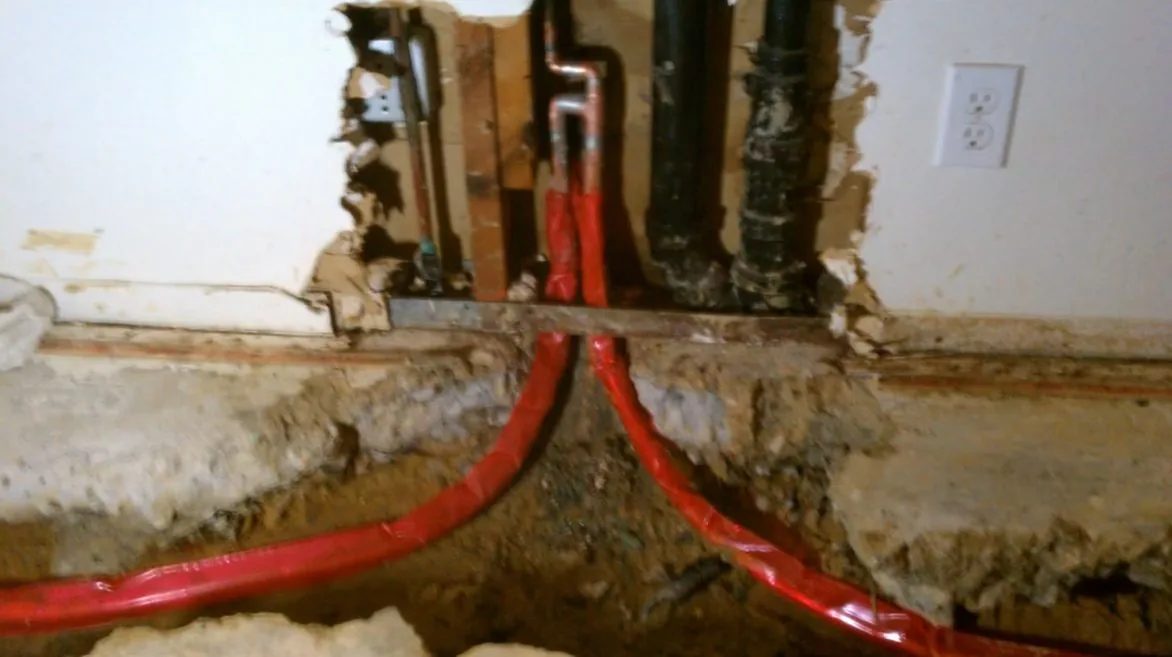 Two red PEX lines running into a channel cut in a concrete floor beneath a wall that has been opened.