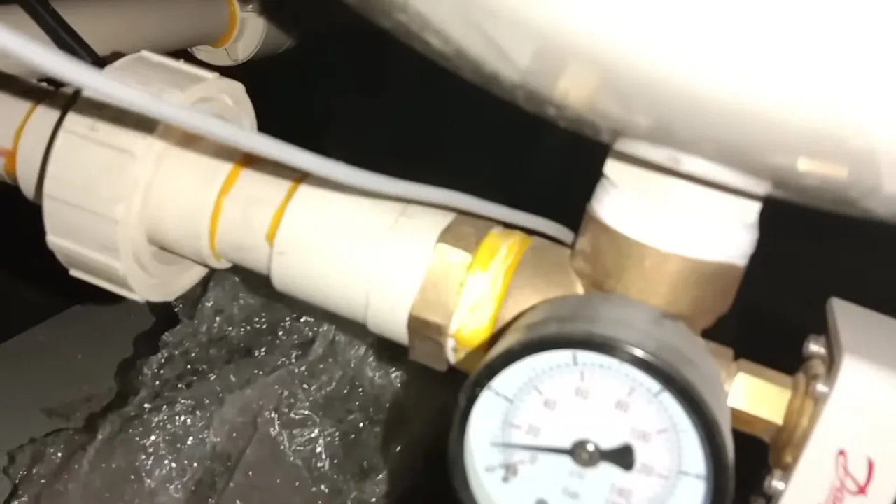 Round pressure gauge on a brass tee beside a white pipe union, with plastic sheeting below.