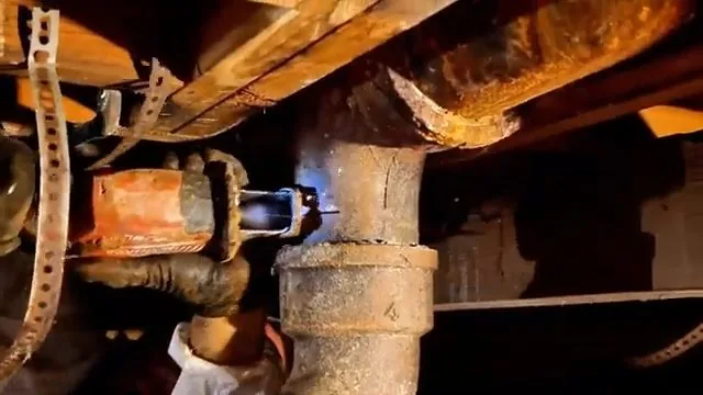 Heavily rusted cast iron drain joint held by a perforated strap beneath wooden floor framing.