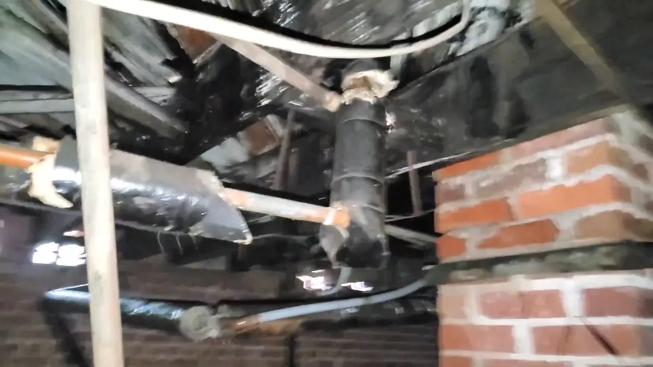 Rusted plumbing joint and pipes beneath a house, beside a brick pier.